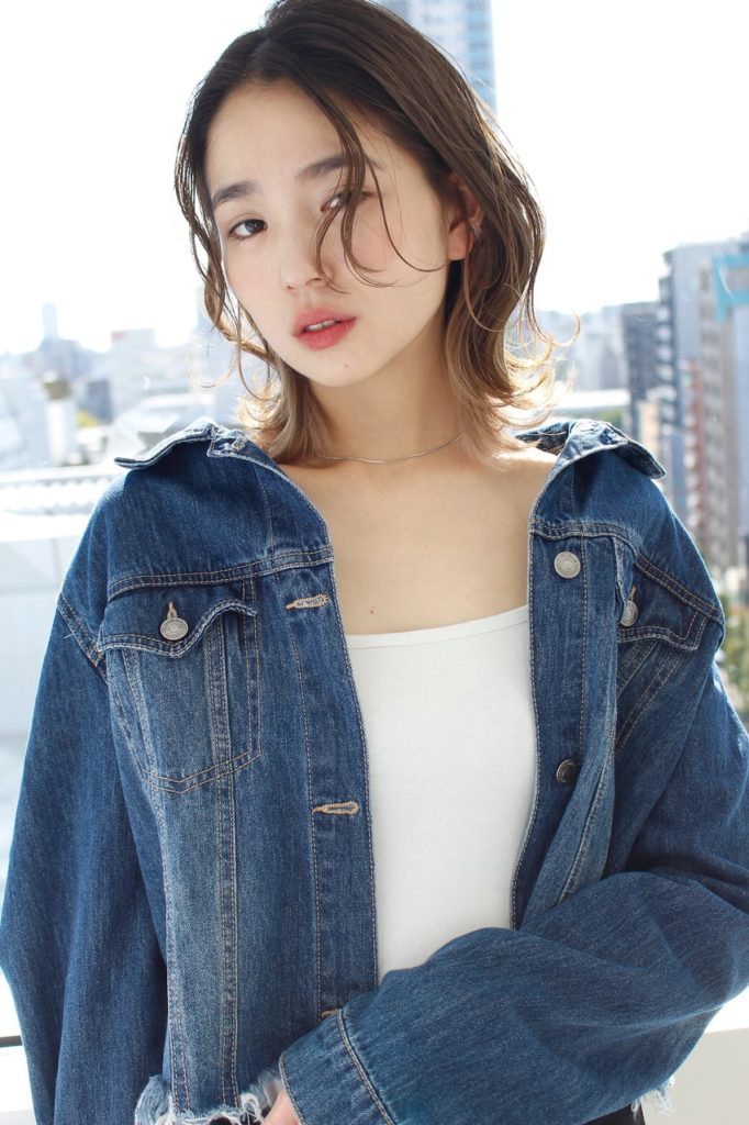都会の街並みをバックに窓際に立つ、センター分けショートヘアの女性の写真